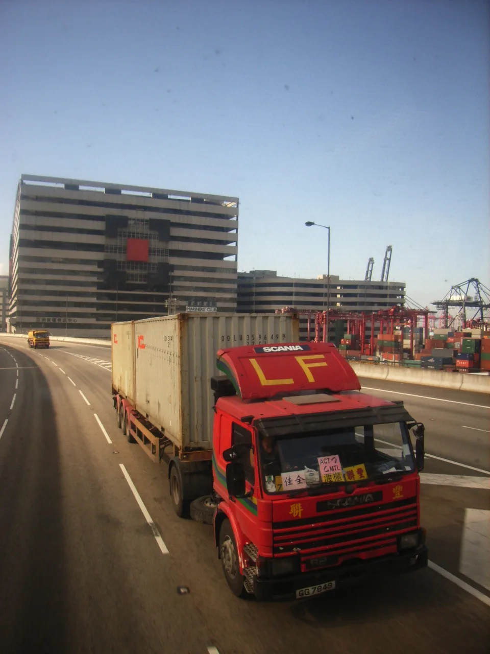 Rode vrachtwagen met zeecontainer op een verhoogde weg, met op de achtergrond een containerterminal met portaalkranen en gestapelde containers.